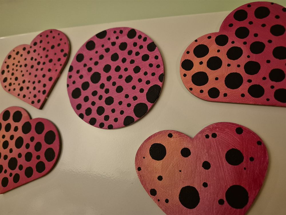 Five painted canvas board cut-outs — four hearts and one circle — in a pink to coral gradient, each covered in irregular black dots, laid out on a white surface.
