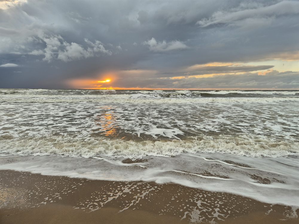 The setting sun breaking through a gap beneath heavy grey rain cloud over the sea, laying a narrow orange stripe across the breaking surf.